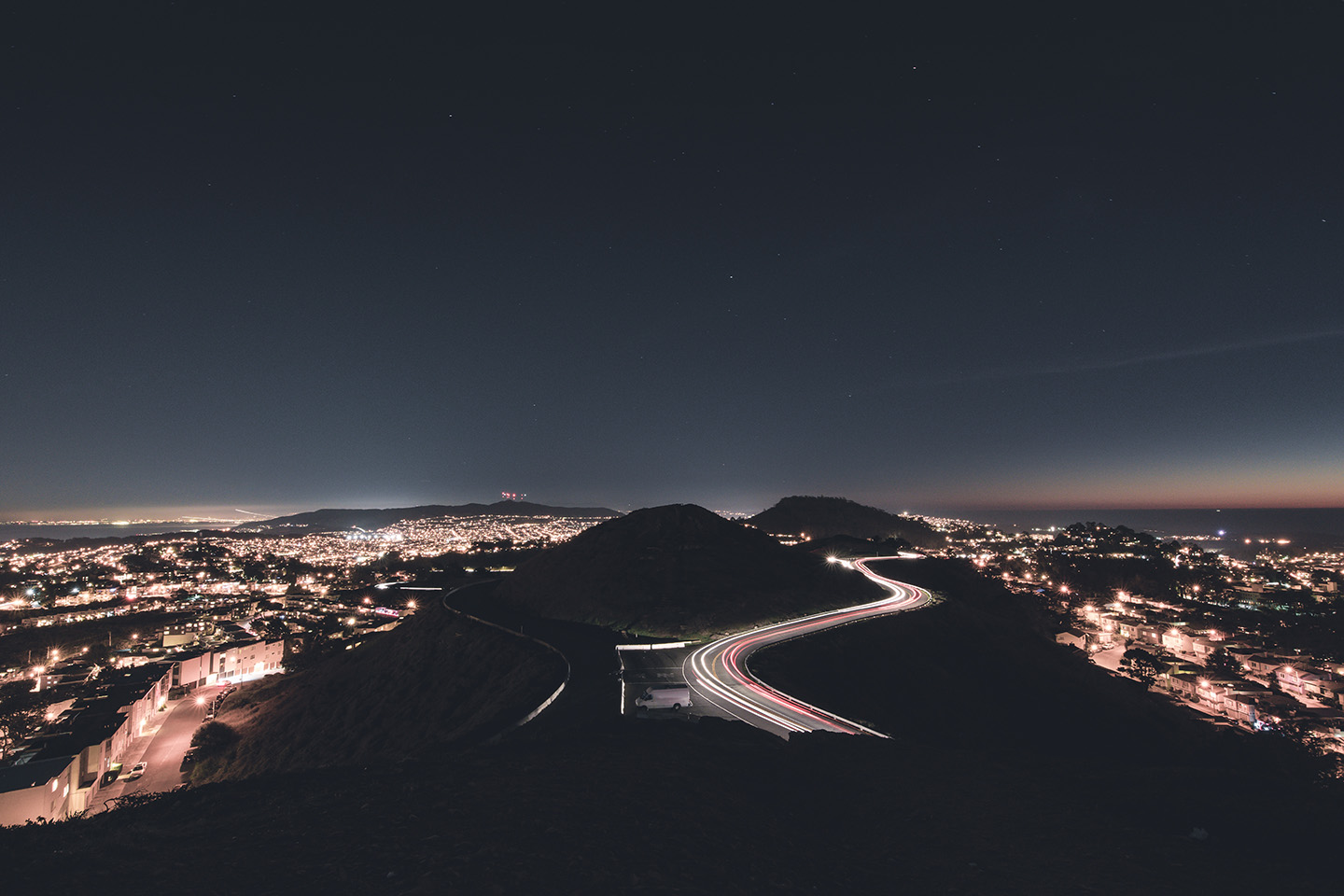 Aerial night view of a city with a winding road showing light trails between dark hills, illuminated neighborhoods in the distance