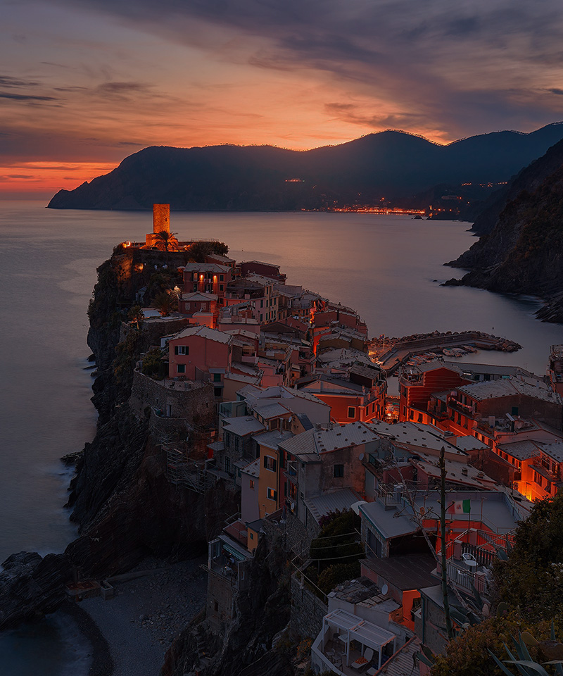 Coastal cliffside village with colorful buildings and a tower at sunset, overlooking calm sea and mountains